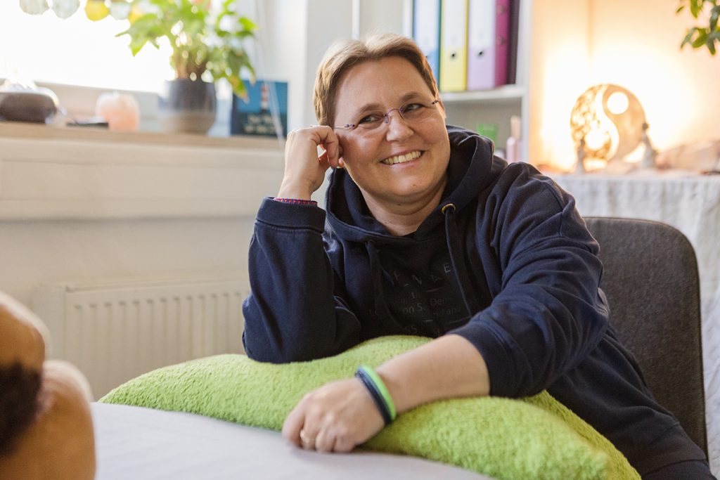 Kontakt mit Kerstin Biß Kerstin Biß sitzt lächelnd an einem Tisch in ihren Räumen.