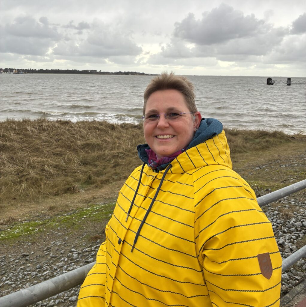 Tine steht am Meer in einer gelben Jacke und blickt ruhig in die Kamera – ein Moment der Klarheit und Neuorientierung