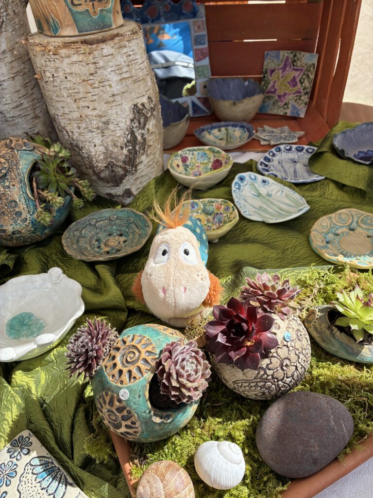 Finnea im Farbenmeer Hörschnecke sitzt zwischen handgemachter Keramik, kleinen Pflanzen und Naturmaterialien auf einem dekorierten Marktstand.