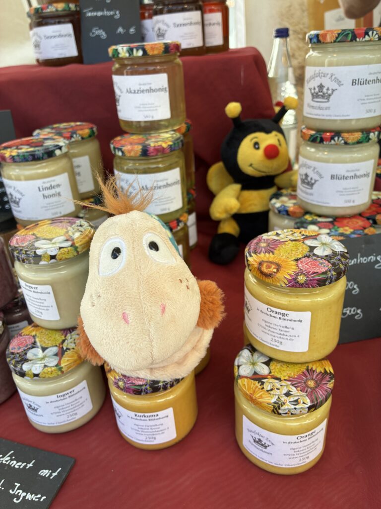 Finnea zu Besuch bei Willy Plüsch-Hörschnecke sitzt zwischen verschiedenen Honiggläsern auf einem Marktstand, im Hintergrund ein kleines Bienen-Kuscheltier.