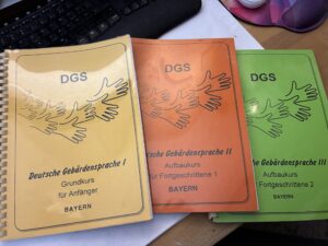 Drei ältere Kursordner zur Deutschen Gebärdensprache für Anfänger und Fortgeschrittene liegen auf einem Schreibtisch.