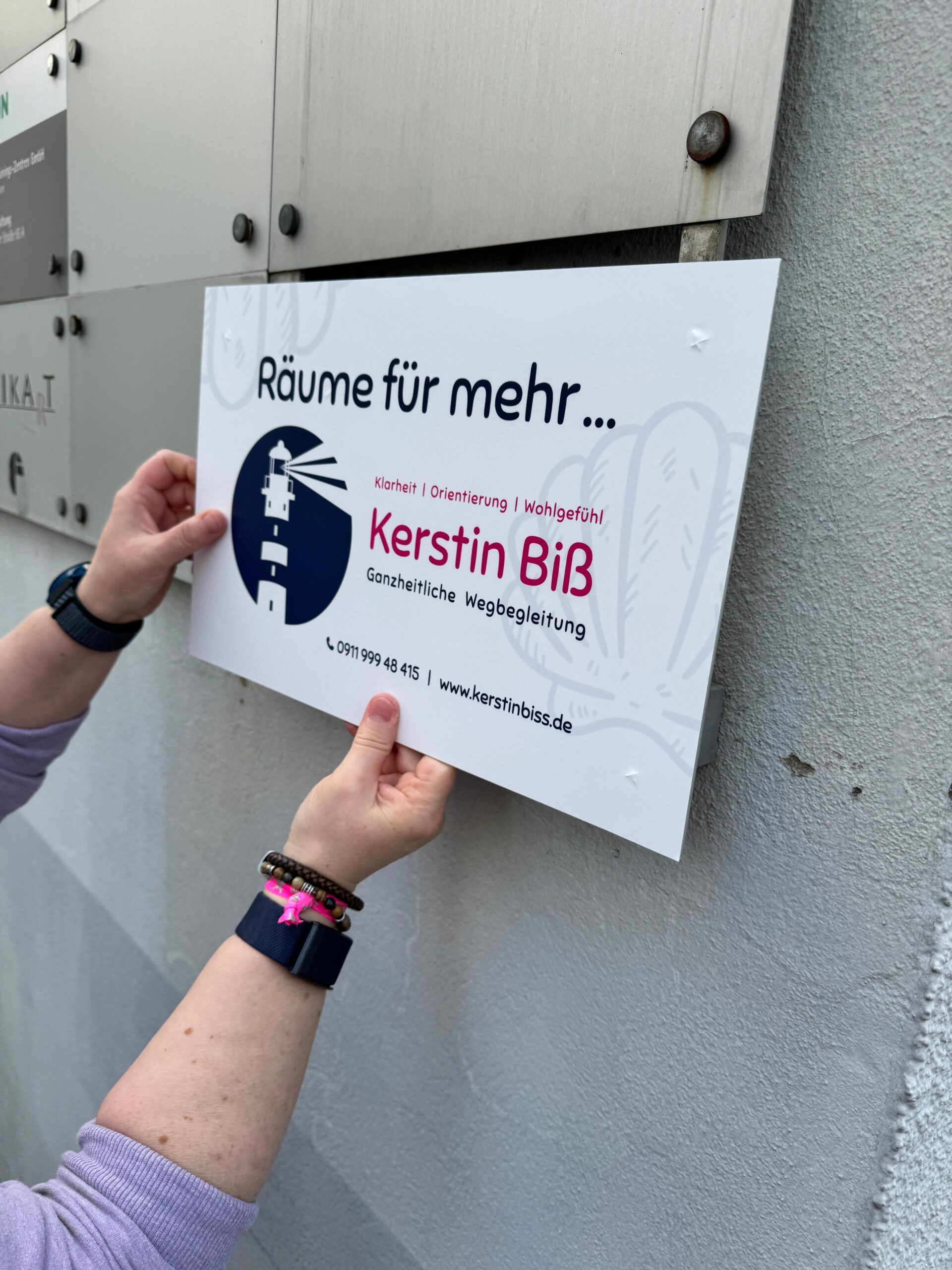 Hände befestigen das neue Firmenschild von Kerstin Biß – Räume für mehr… an der Hauswand.