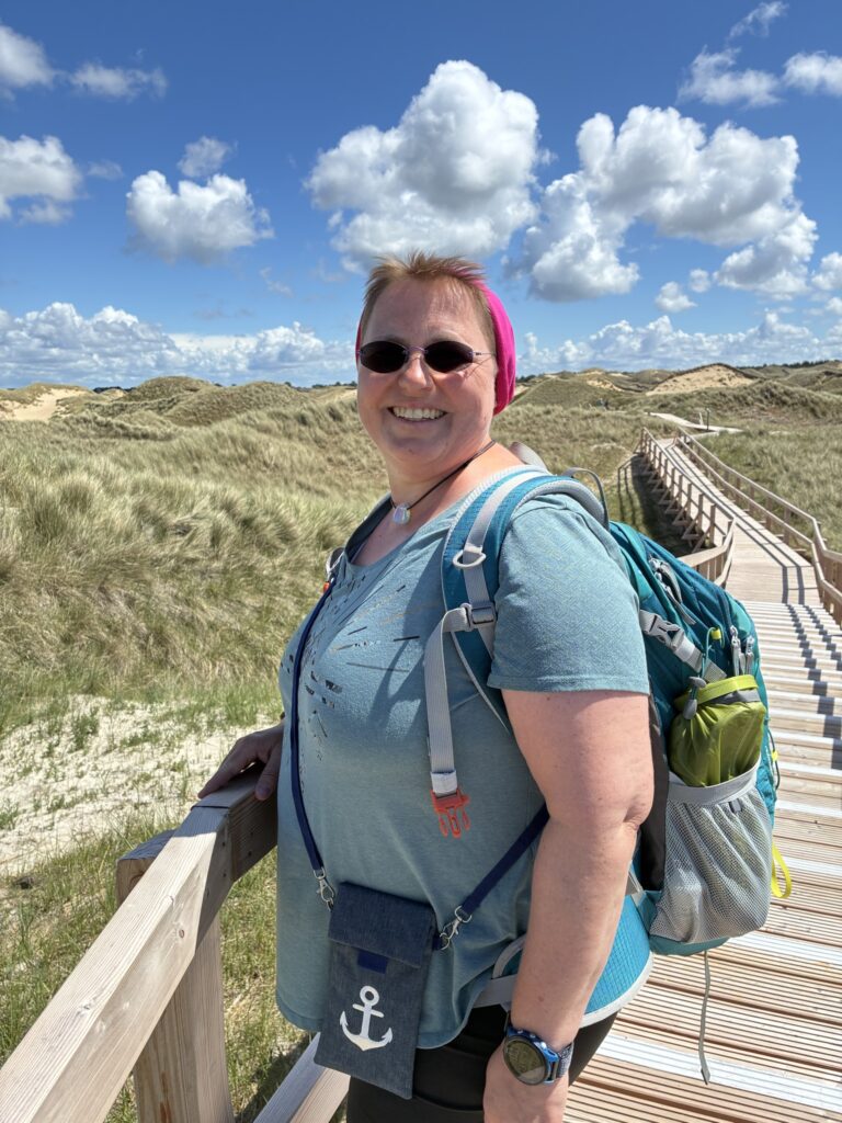 Kerstin Biß steht auf einem Holzsteg zwischen Dünen unter blauem Himmel.