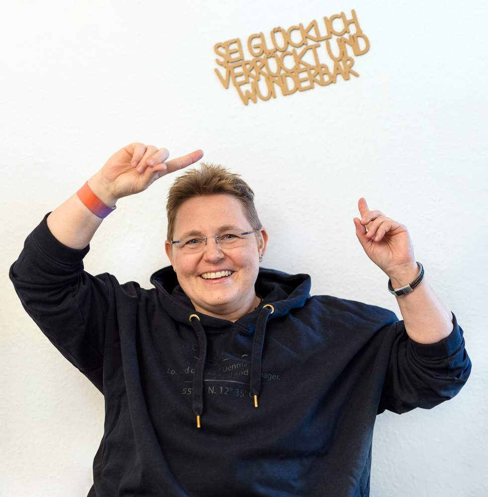 Kerstin Biß lächelt und zeigt mit beiden Händen auf den Schriftzug „Sei glücklich, verrückt und wunderbar“ an der Wand.